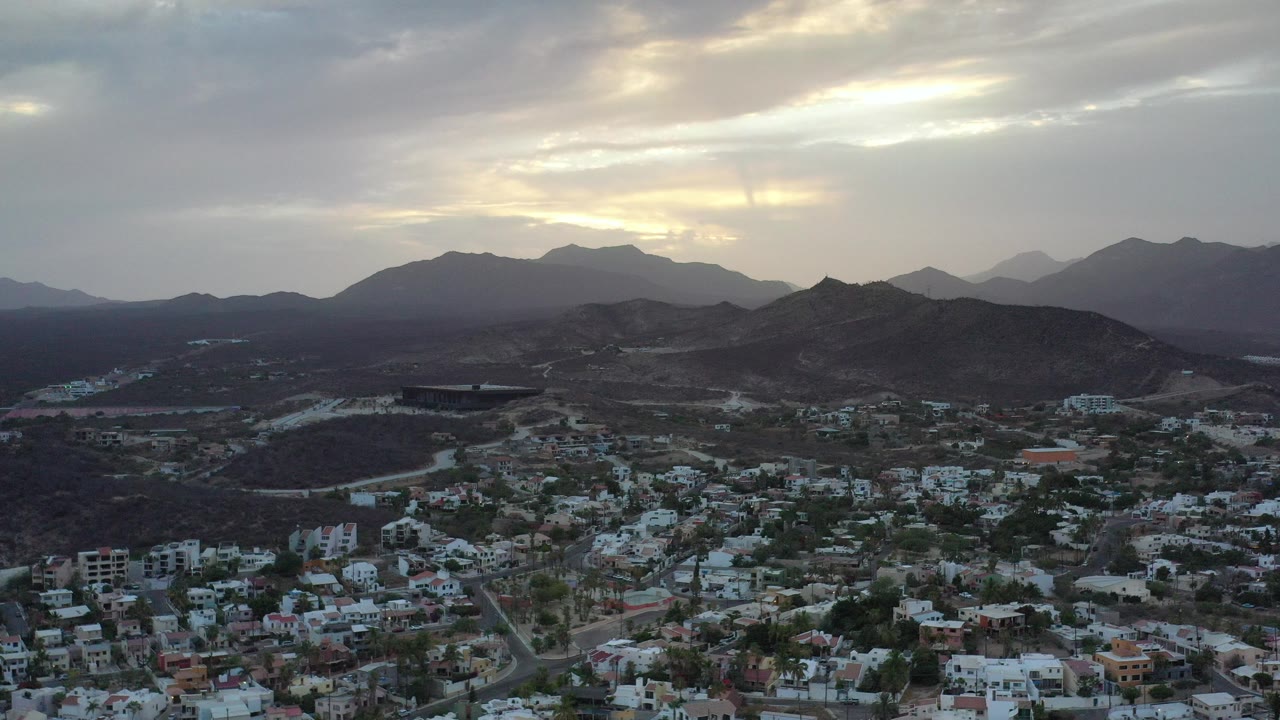 Aerial city view below dark hills and a cloudy sunset sky.