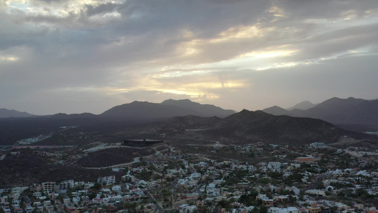 Wide aerial frame over Los Cabos, Mexico, with clustered houses at the bottom, dark hills across the middle, sunlit clouds above, and no visible people.