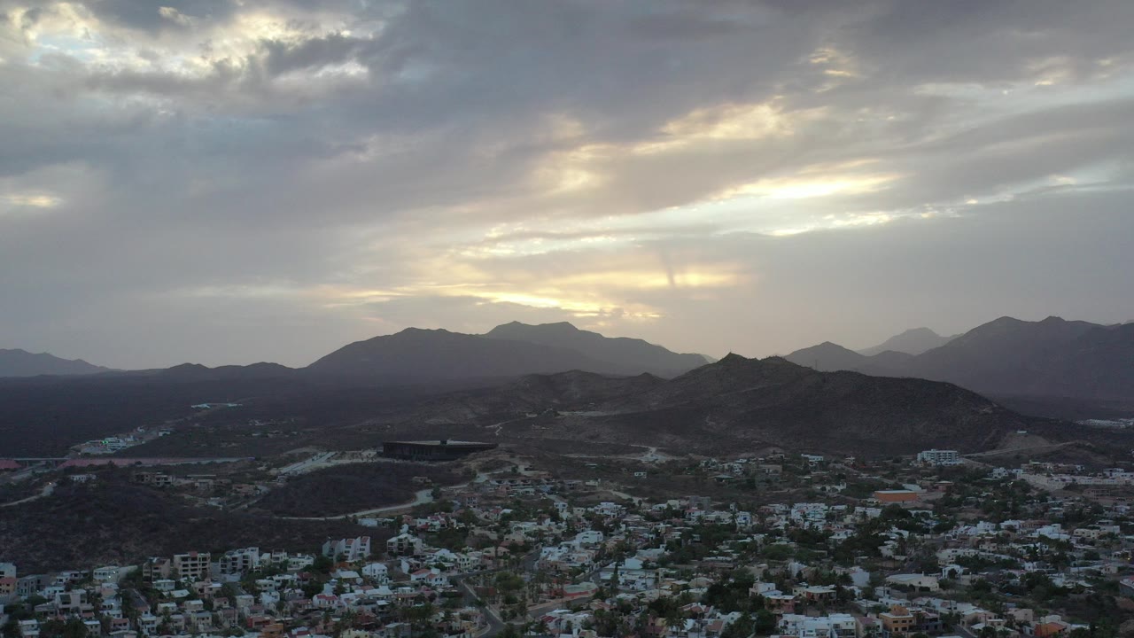 Early frame of Los Cabos, Mexico, showing low white buildings below dark hills and layered mountains under gray sunrise clouds, with no visible people.