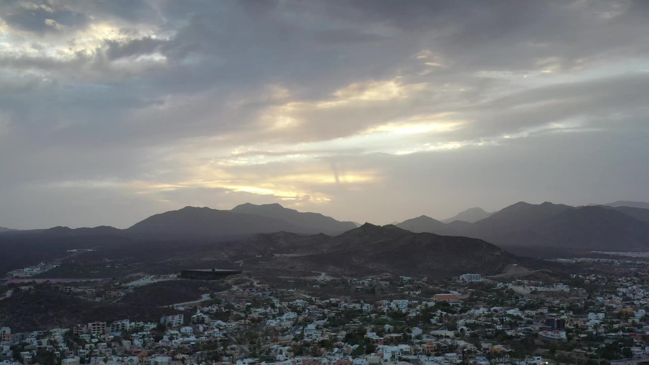 Aerial Los Cabos frame with the city spread across the lower foreground, layered mountain ridges in the distance, muted gray-blue clouds, and no visible people.