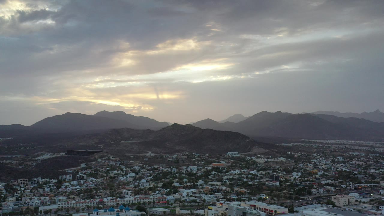 Wide aerial frame over Los Cabos, Mexico, showing town buildings beneath dark hills and layered gray clouds lit by yellow sunset, with no visible people.