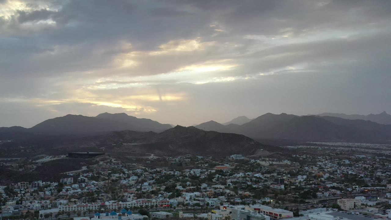Aerial view in Los Cabos, Mexico, with dense buildings in the lower frame, a dark mountain ridge across the center, and yellow light breaking through gray clouds, with no visible people.