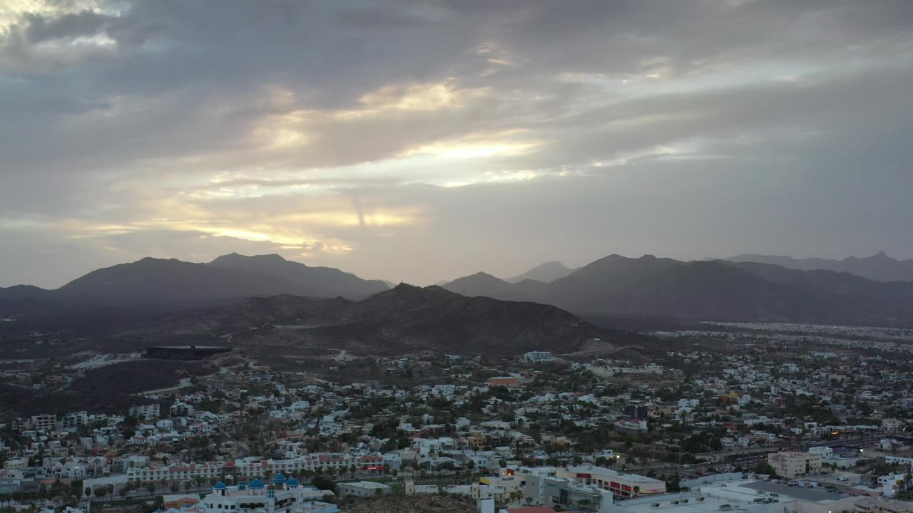 Town rooftops, low hills, and distant mountains under a bright cloud break.