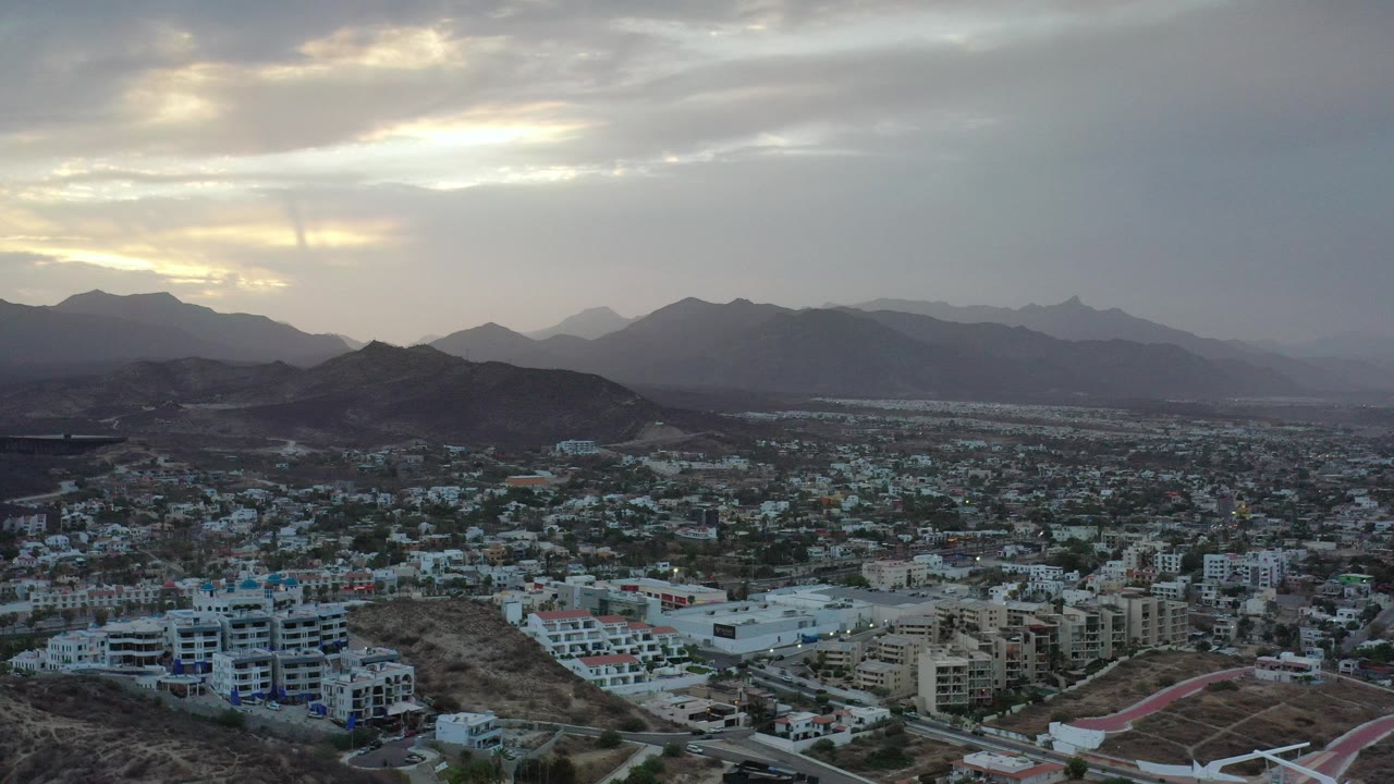 Aerial view of city neighborhoods below dark hills and cloudy mountains.