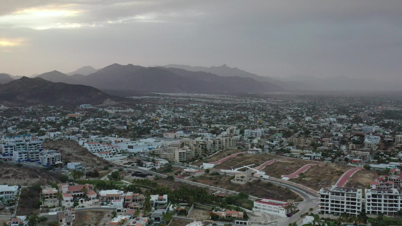 Aerial view of houses, roads, hills, and mountains under gray clouds.
