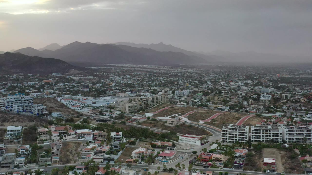 Elevated Los Cabos city view with apartment blocks and houses in the foreground, a broad urban grid in the distance, hazy mountains, gray clouds, and no visible people.
