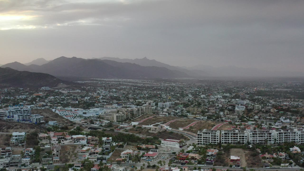 Wide aerial view of city buildings beneath gray clouds and distant mountains.