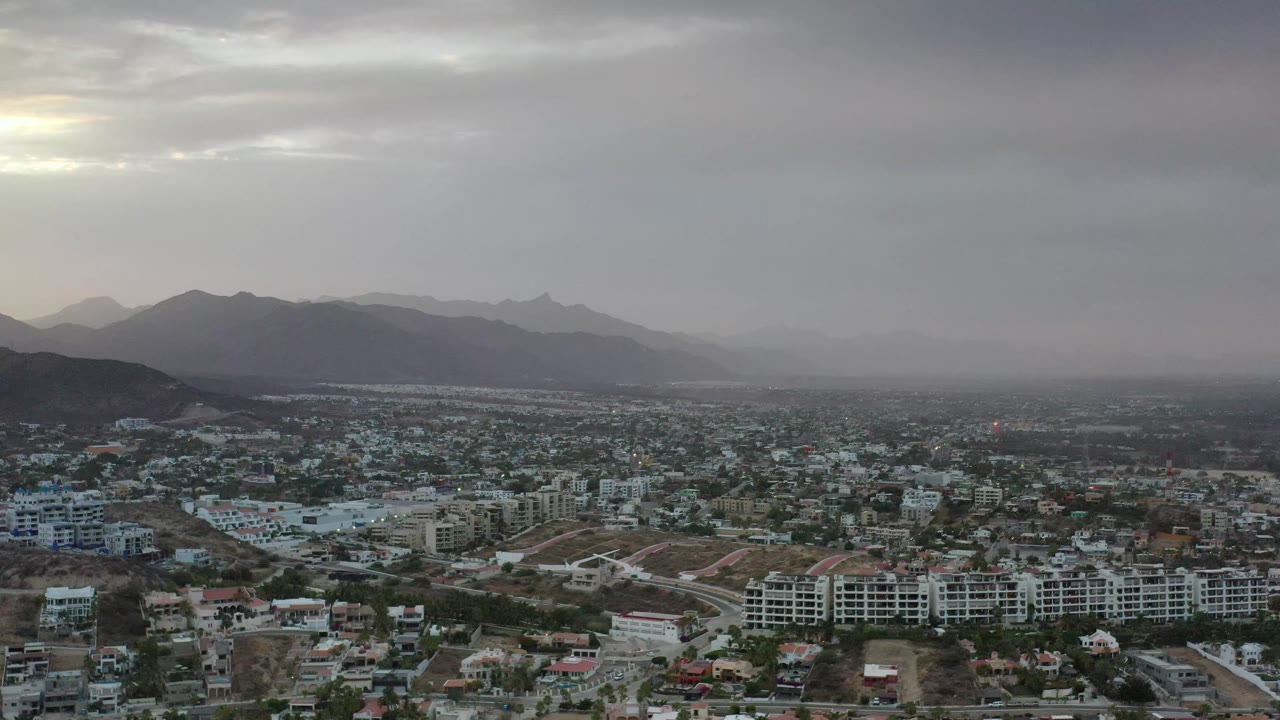 Aerial view across Los Cabos, Mexico, showing clustered low-rise development, pale roads, dark foothills, soft gray sky, and no visible people.