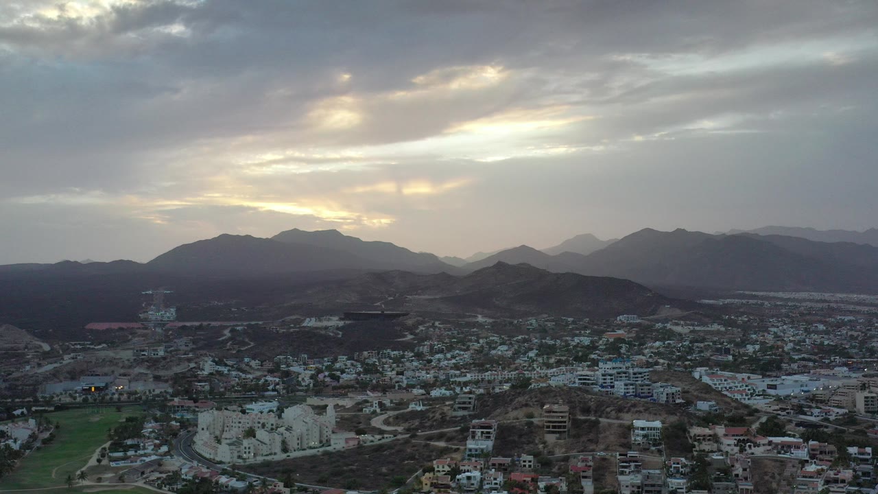 Elevated town view below dark mountains and a cloudy sunset sky.
