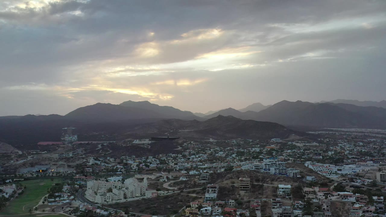 A wide Los Cabos, Mexico view shows clustered rooftops, winding roads, dark hills, and pale sunset light filtering through clouds, with no visible people.