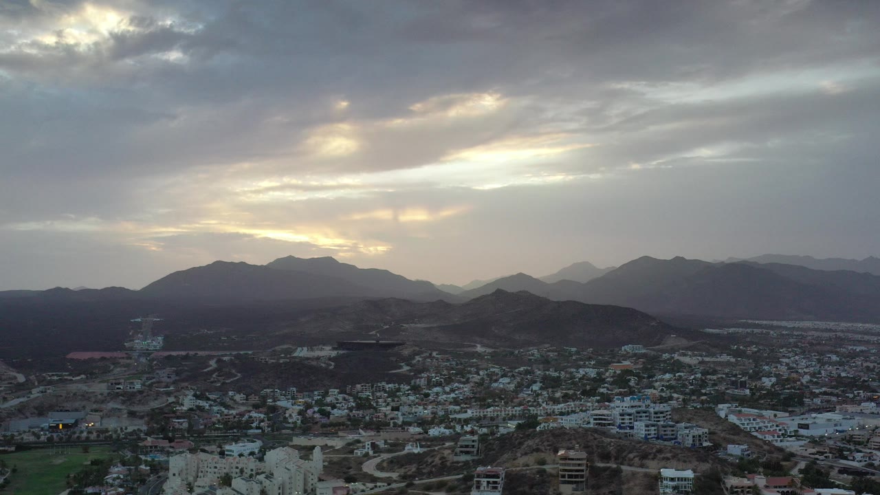 Opening frame of Los Cabos, Mexico with a low townscape below dark mountain ridges and pale gold light breaking through dense clouds, no visible people.