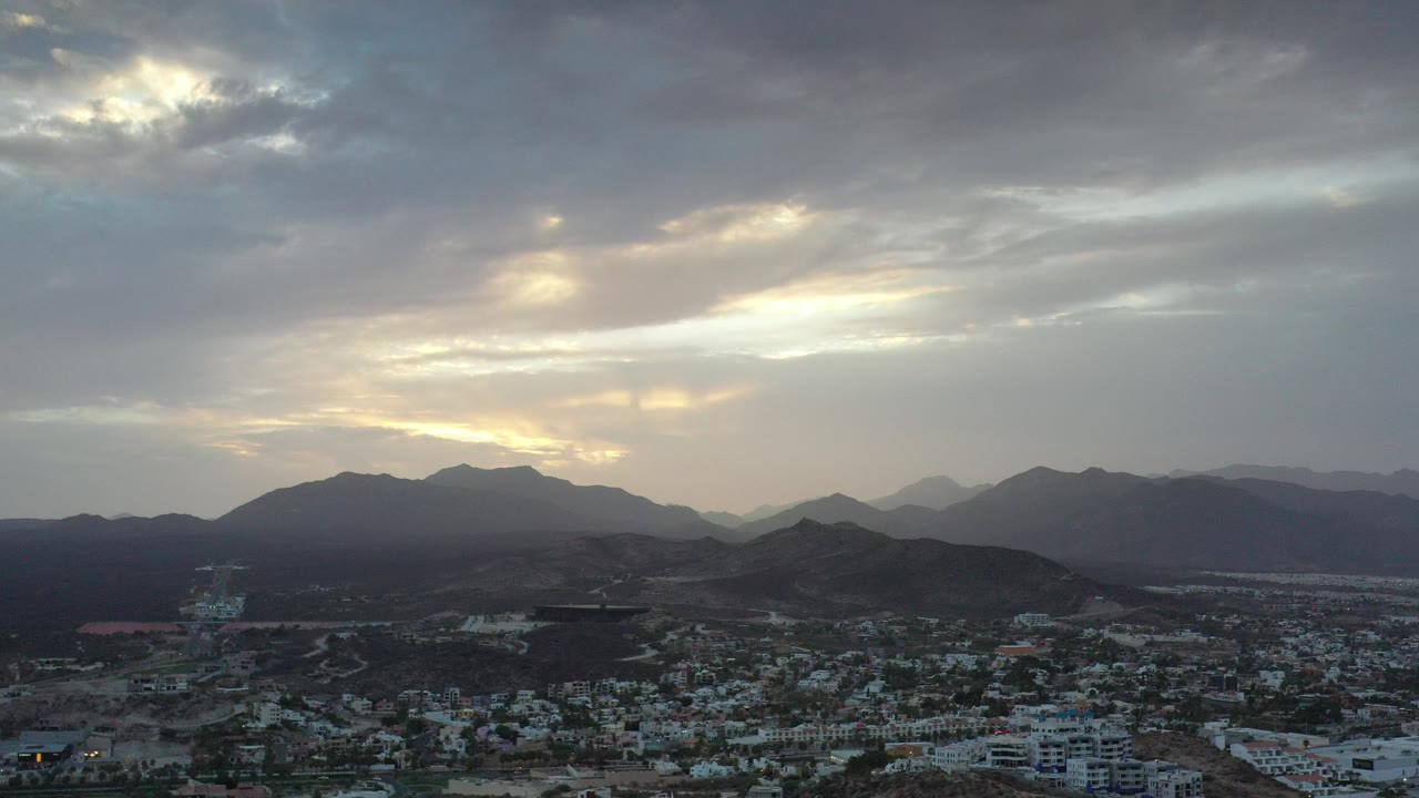 Aerial view of city buildings below dark hills and sunlit gray clouds.