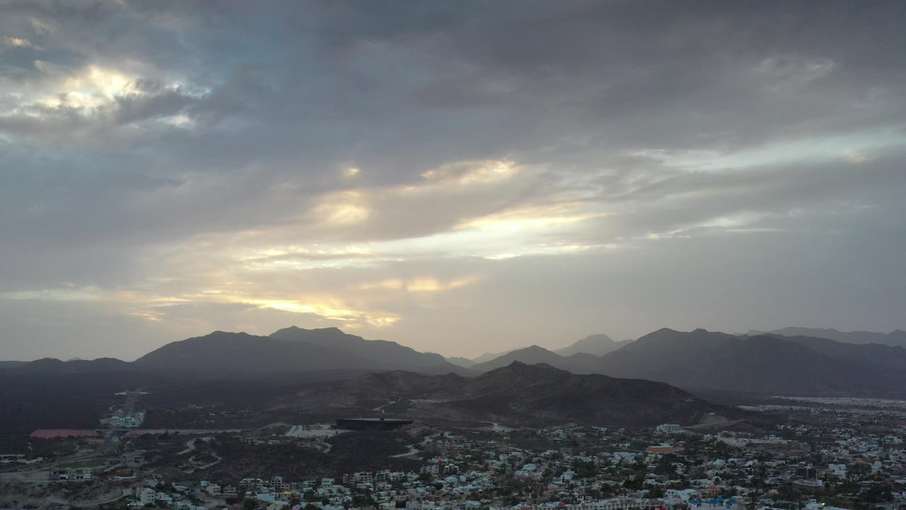 Opening frame of Los Cabos, Mexico, showing a low city area beneath dark mountain ridges, heavy gray clouds, warm sunlight near the horizon, and no visible people.