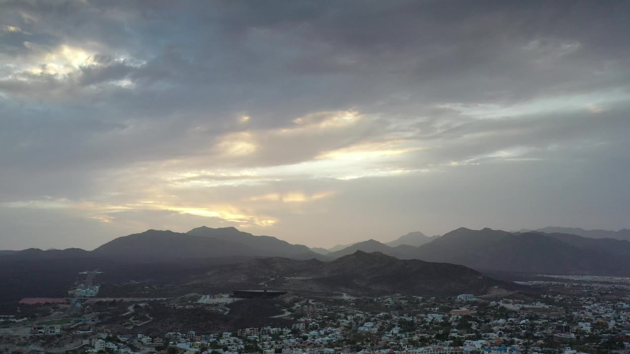 Cloudy sunset sky over mountain ridges and a distant coastal city.