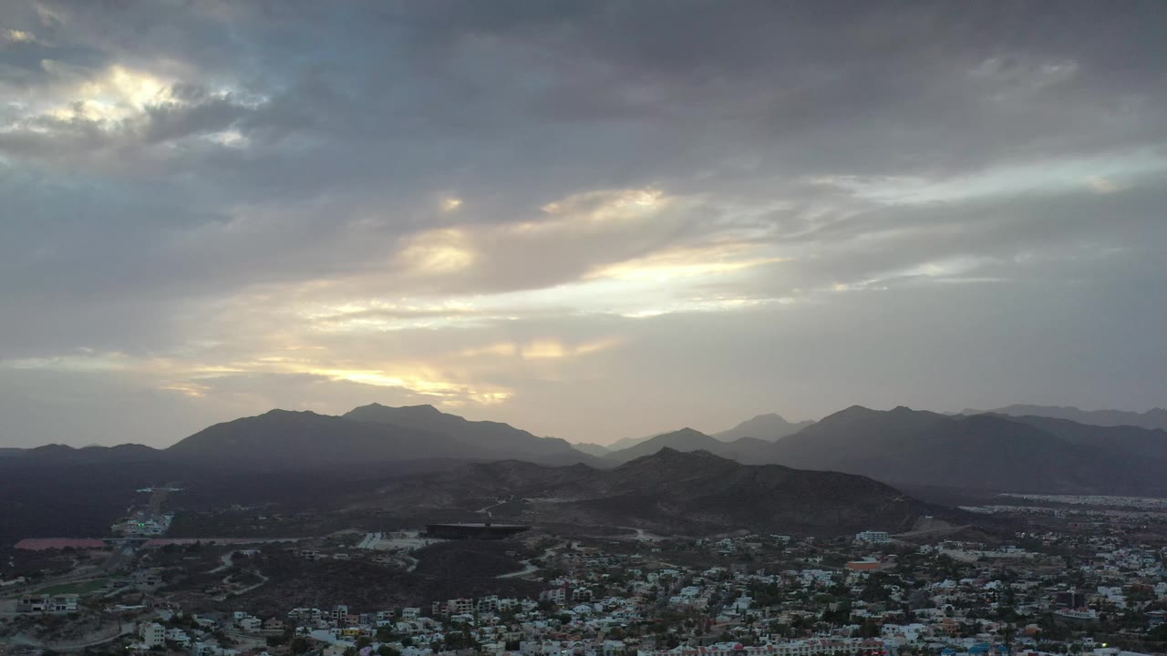 Aerial Los Cabos landscape with a bright opening in the cloud cover, silhouetted mountain layers across the horizon, low hills, distant town buildings, and no visible people.