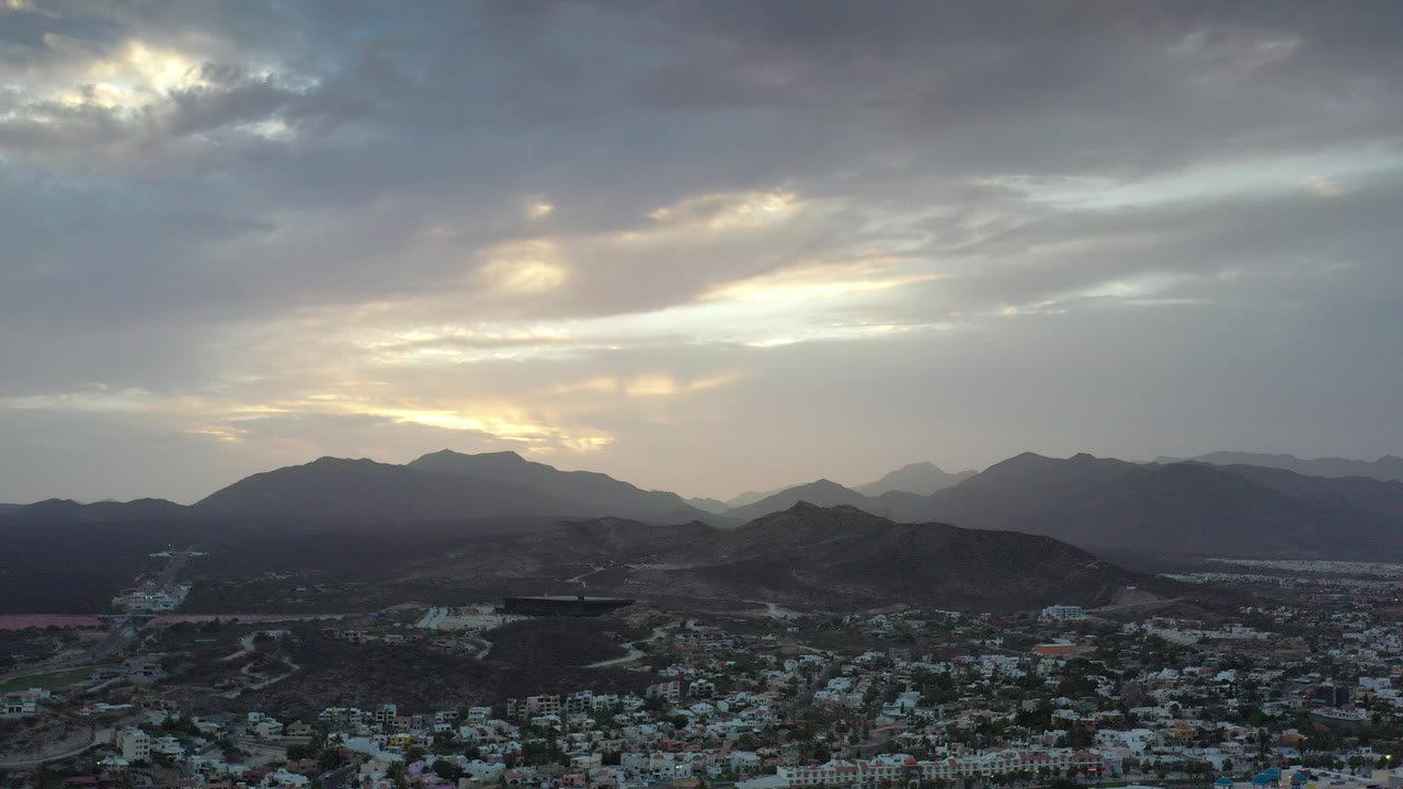 Wide aerial view in Los Cabos, Mexico, showing a cloudy sunrise over dark mountain ridges, desert hills, and a town in the foreground with no visible people.