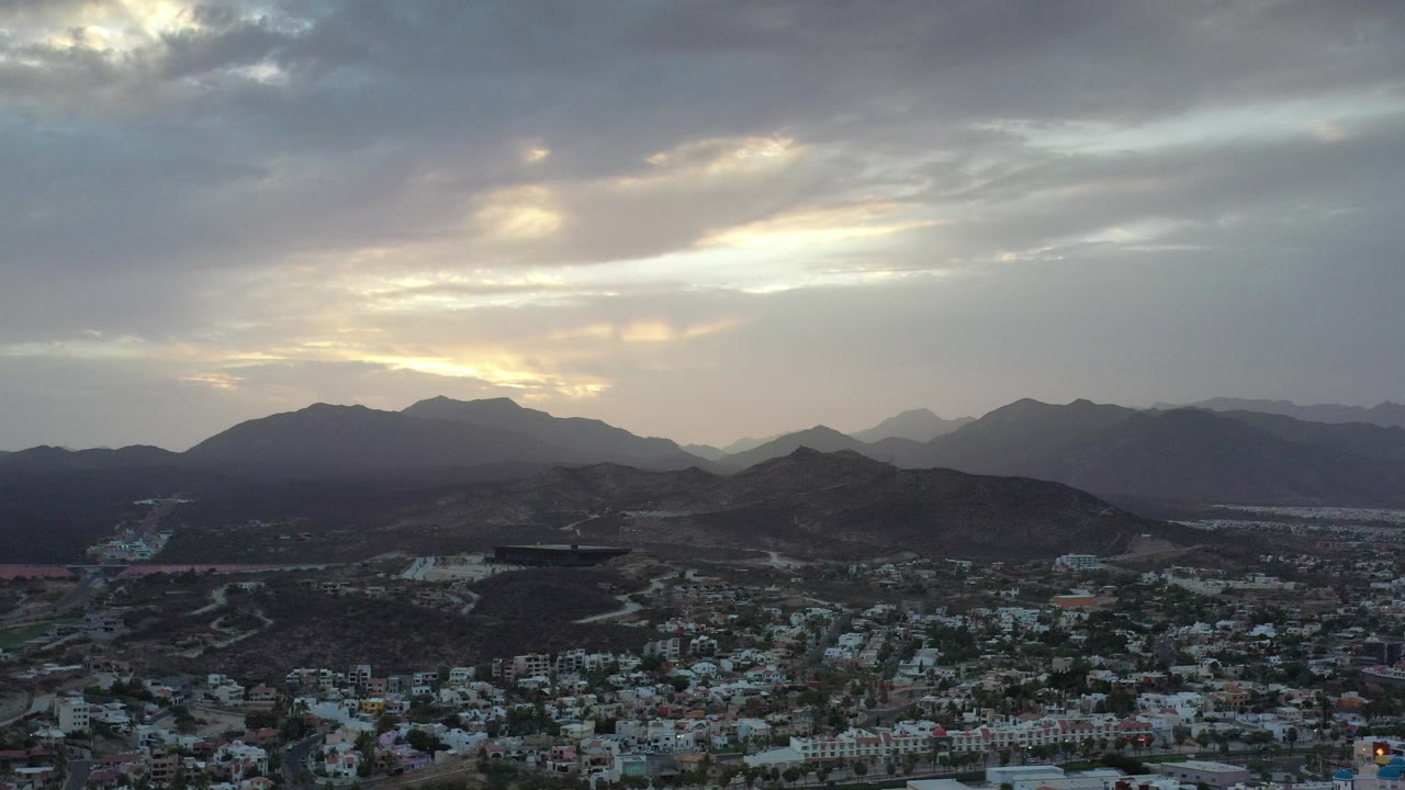 Opening aerial frame over Los Cabos, Mexico, with town rooftops at the bottom, dark hills across the middle, layered mountains in the distance, and no visible people.