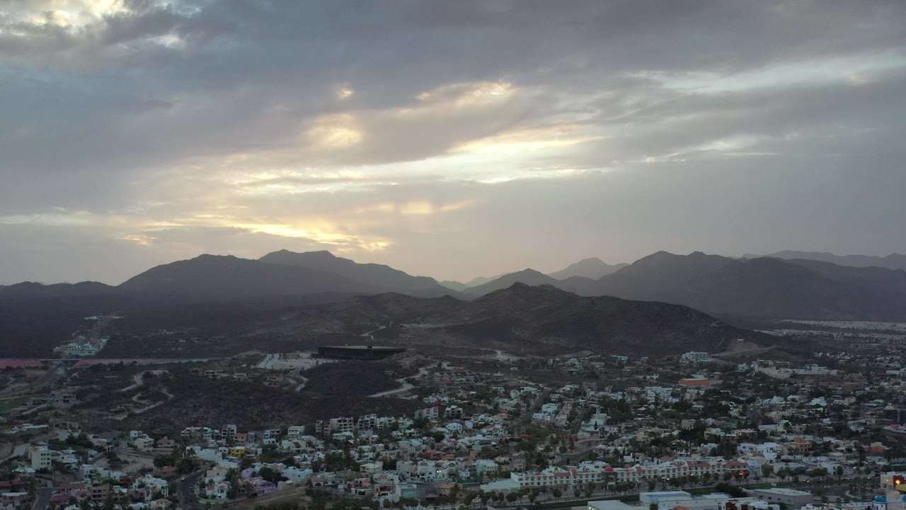 Town and arid hills beneath layered mountains and a bright cloudy sky.