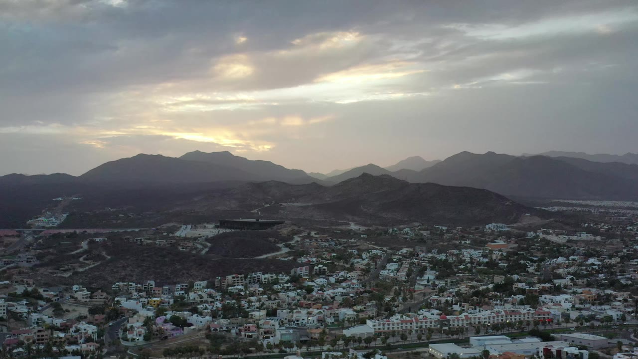 A high view over Los Cabos, Mexico, shows the city spread across the lower frame, rounded hills in the middle distance, a brighter opening in the evening clouds, and no visible people.