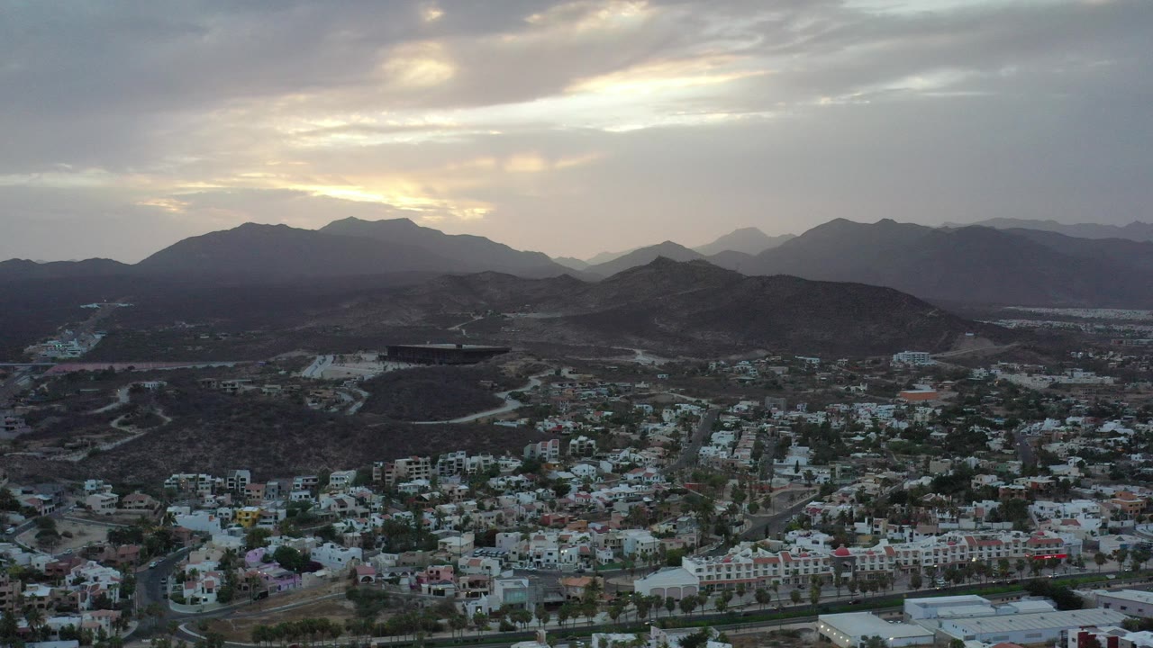 Wide aerial view over Los Cabos, Mexico, with low-rise buildings below dark desert hills and layered mountains under a cloudy sunrise sky, no visible people.