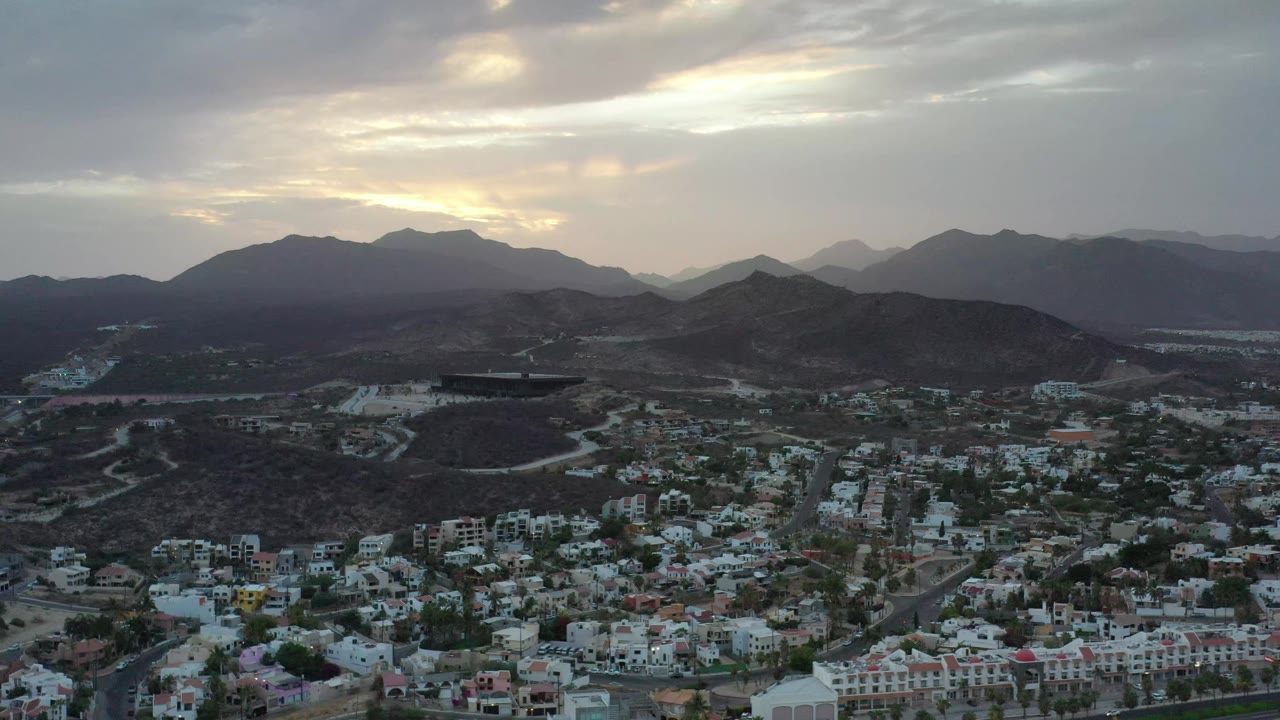 Early frame of the Los Cabos aerial shows white buildings and roads spread across the lower view, with darker hills and hazy mountains under a gray cloud deck, and no visible people.