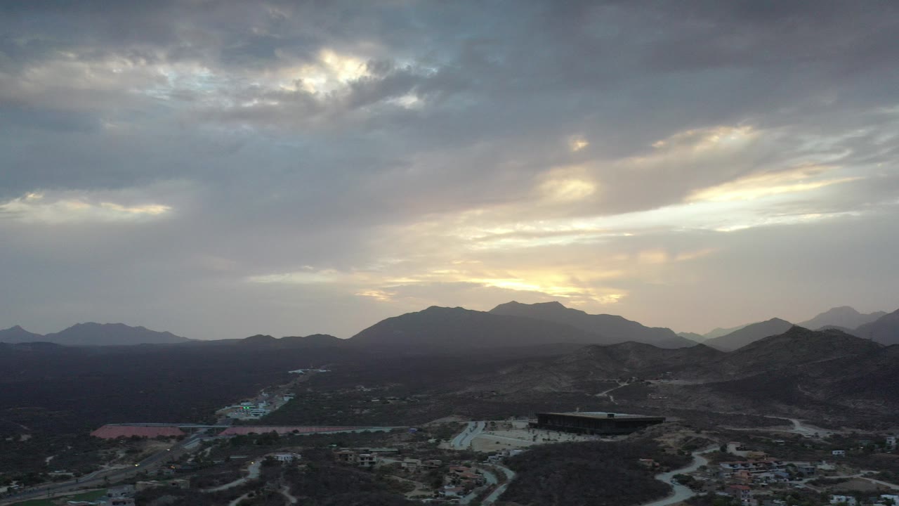 Wide aerial view over Los Cabos, Mexico, with winding roads, dark desert hills, layered clouds, and no visible people.