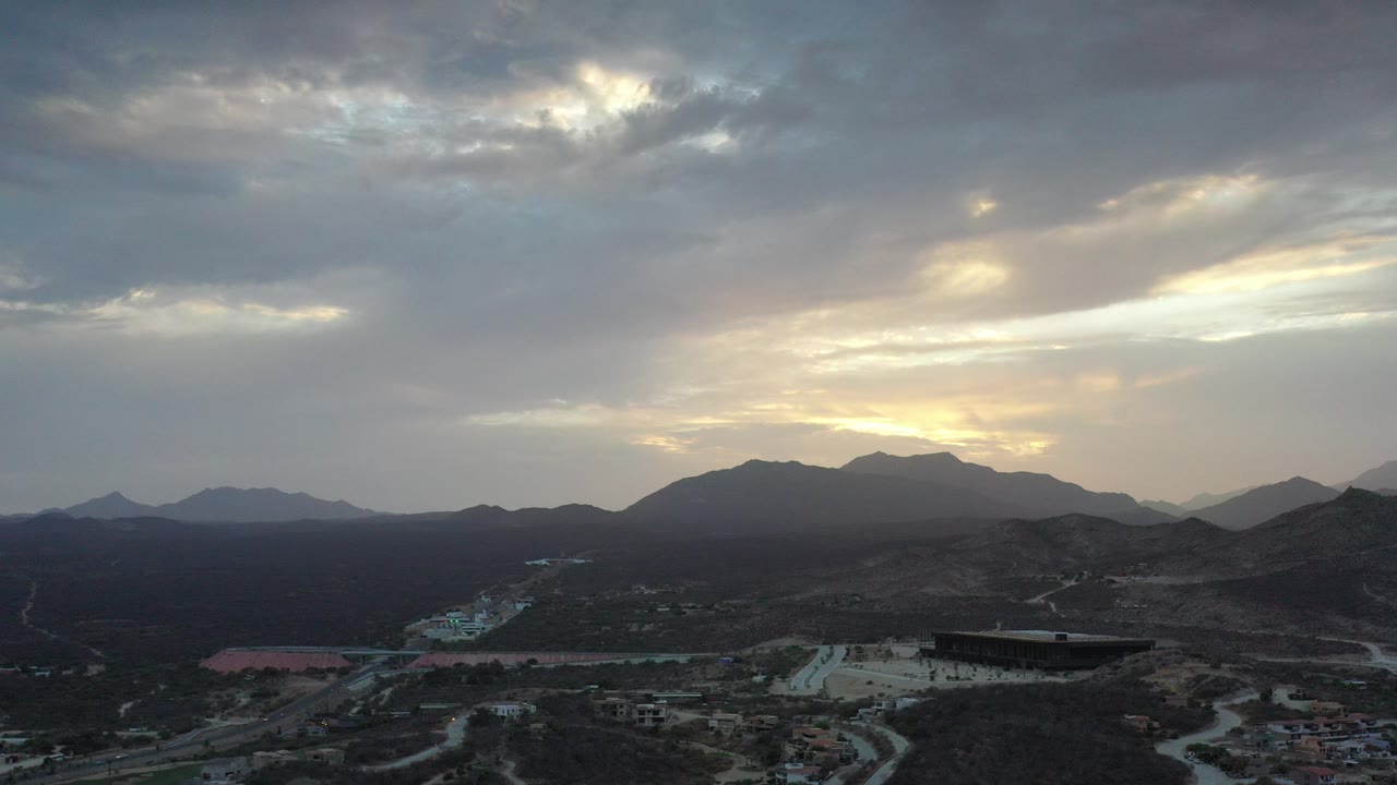 Opening frame of Los Cabos, Mexico with dark mountain ridges, pale roads, scattered low buildings, layered clouds, warm horizon light, and no visible people.