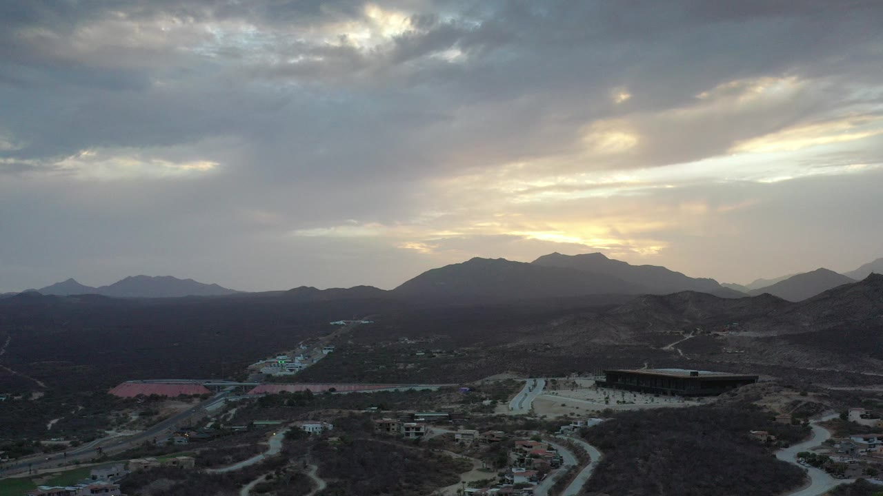 Wide aerial view over Los Cabos, Mexico with dark mountain ridges, scattered roads and buildings below, warm cloud-filtered light, and no visible people.