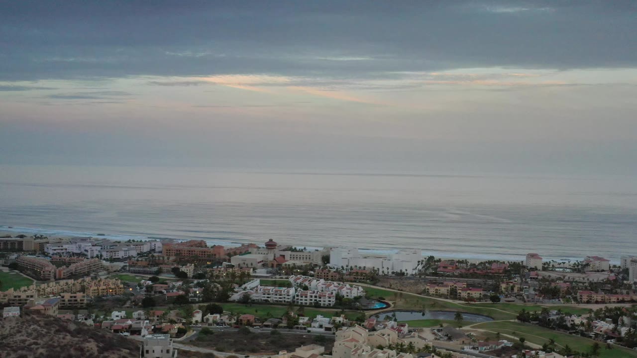 High aerial frame in Los Cabos showing low-rise coastal architecture, pale surf lines, muted ocean water, and layered evening clouds with no visible people.