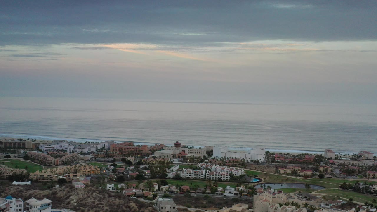 Opening frame of Los Cabos, Mexico from an elevated view, showing coastal buildings, palms, rocky foreground, ocean bands, layered clouds, and no visible people.