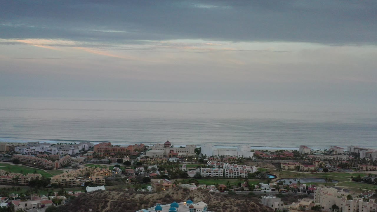 Opening aerial frame over Los Cabos, Mexico with rocky foreground, low-rise coastal buildings, muted ocean water, layered clouds, and no visible people.