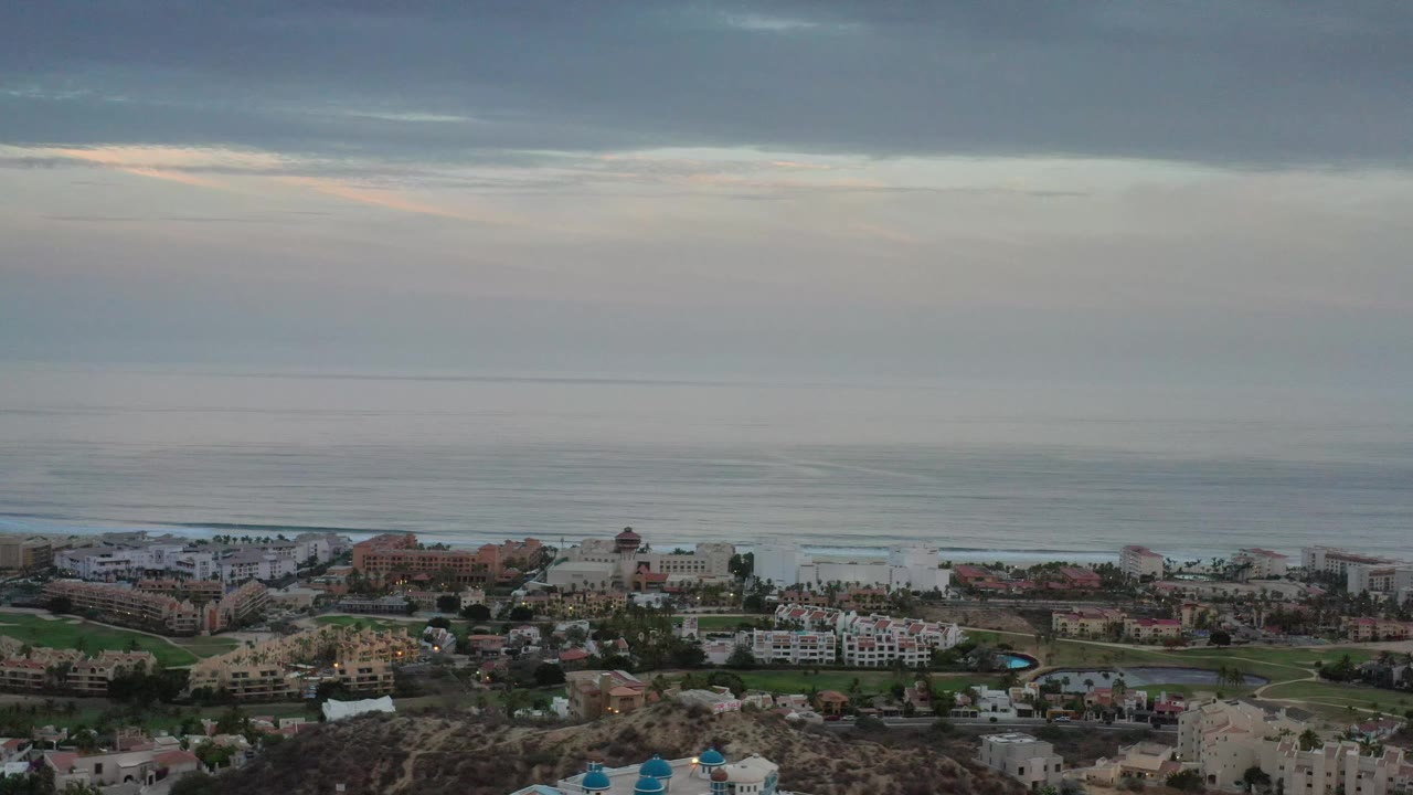 Aerial view in Los Cabos, Mexico showing resort-style buildings, green grounds, rocky terrain at the bottom edge, a flat ocean horizon, and no visible people.