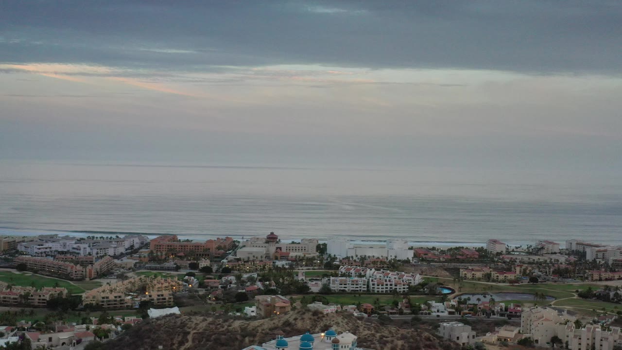 Wide aerial coastline with buildings, palms, and calm ocean water.