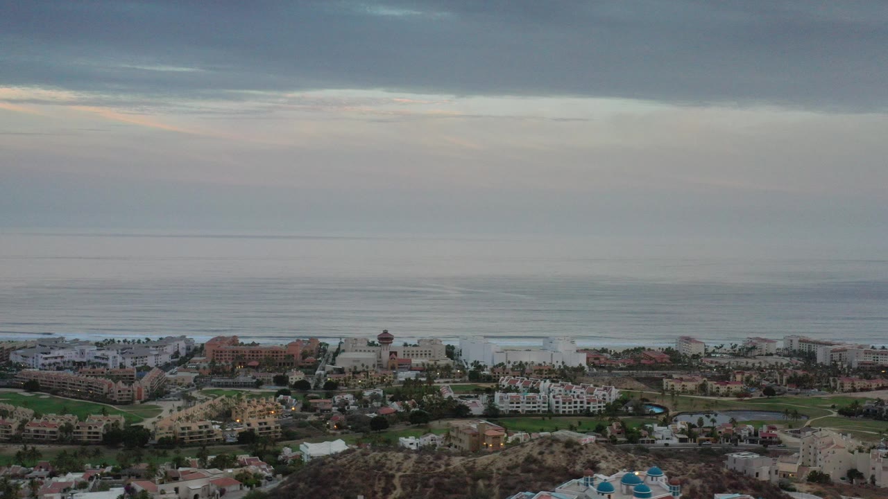 Opening frame of Los Cabos, Mexico from a high vantage, showing resort-style buildings and greenery below a broad gray-blue ocean and clouded sky, with no visible people.