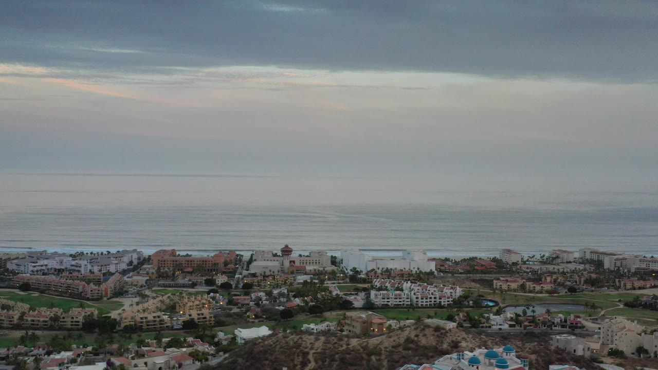A wide Los Cabos view shows the ocean horizon above clustered light-colored buildings, palms, and landscaped grounds, with no visible people.