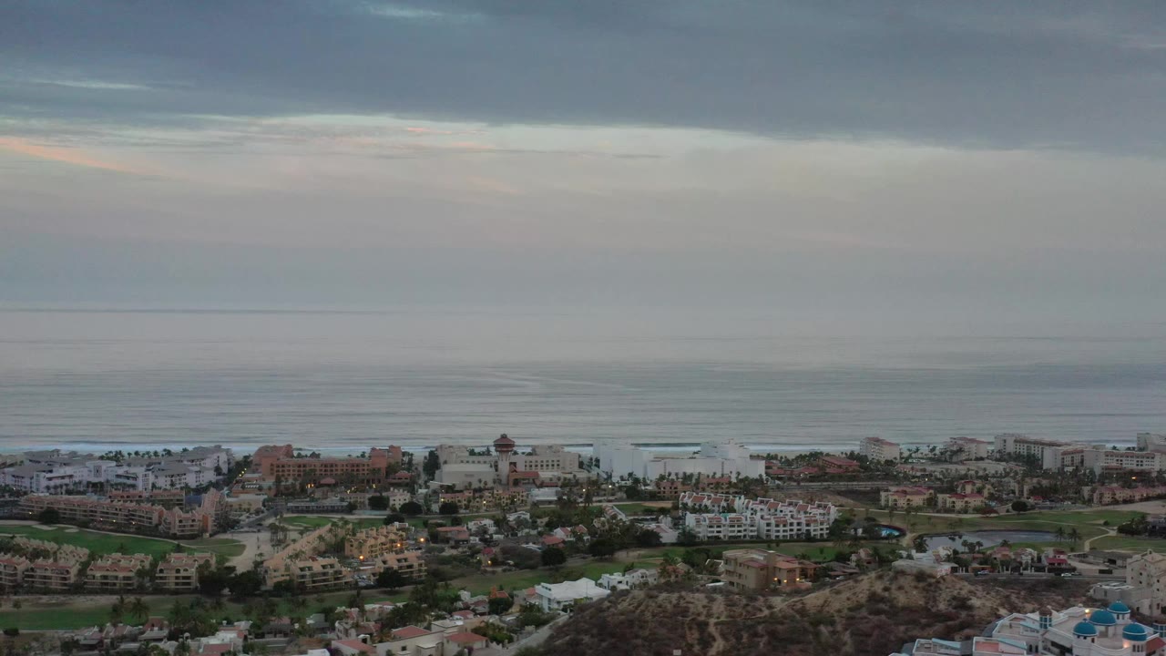 Los Cabos coastal panorama with low surf lines across the sea, clustered shoreline buildings, green patches, and no visible people.