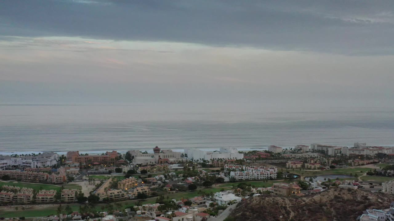 Los Cabos aerial frame showing white and tan buildings across the lower third, calm ocean beyond them, a dark cloud ceiling, and no visible people.