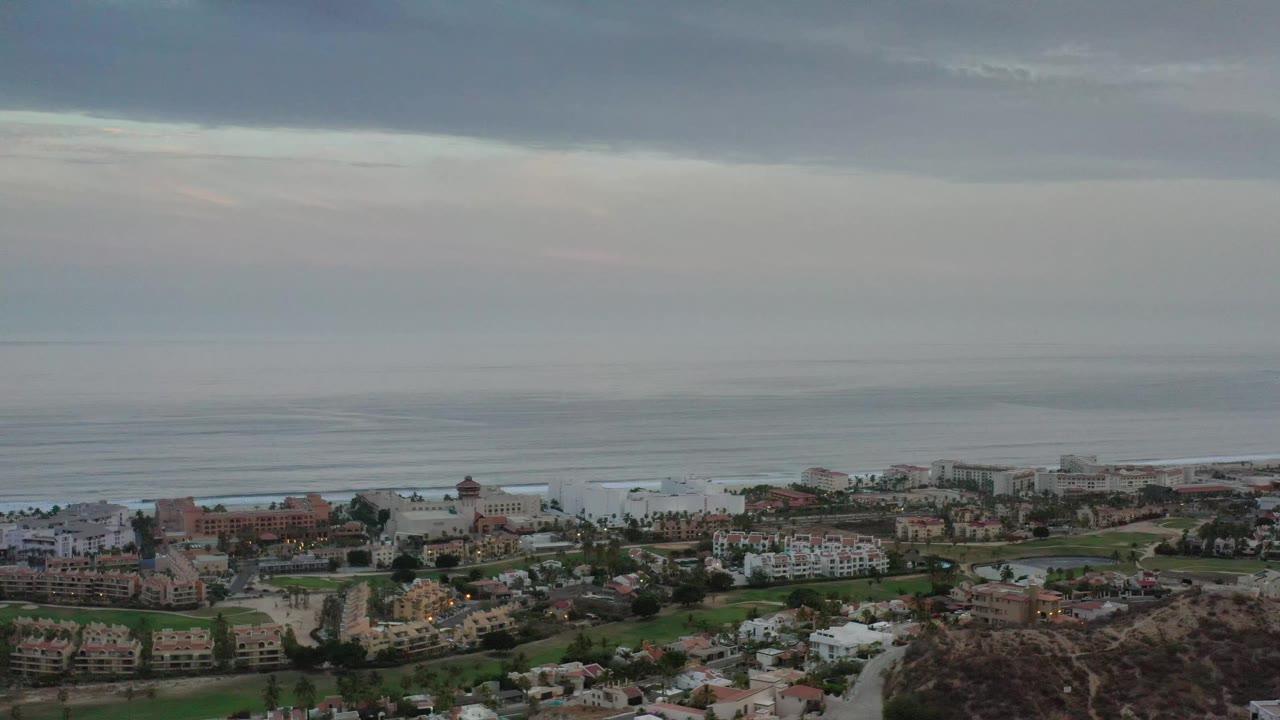 Elevated Los Cabos view with clustered homes and roads in the lower frame, flat ocean water beyond, and no visible people.