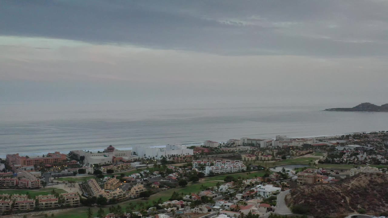 Los Cabos coastal neighborhood seen from above, with clustered buildings along the lower frame, a calm sea horizon, distant offshore landforms, and no visible people.