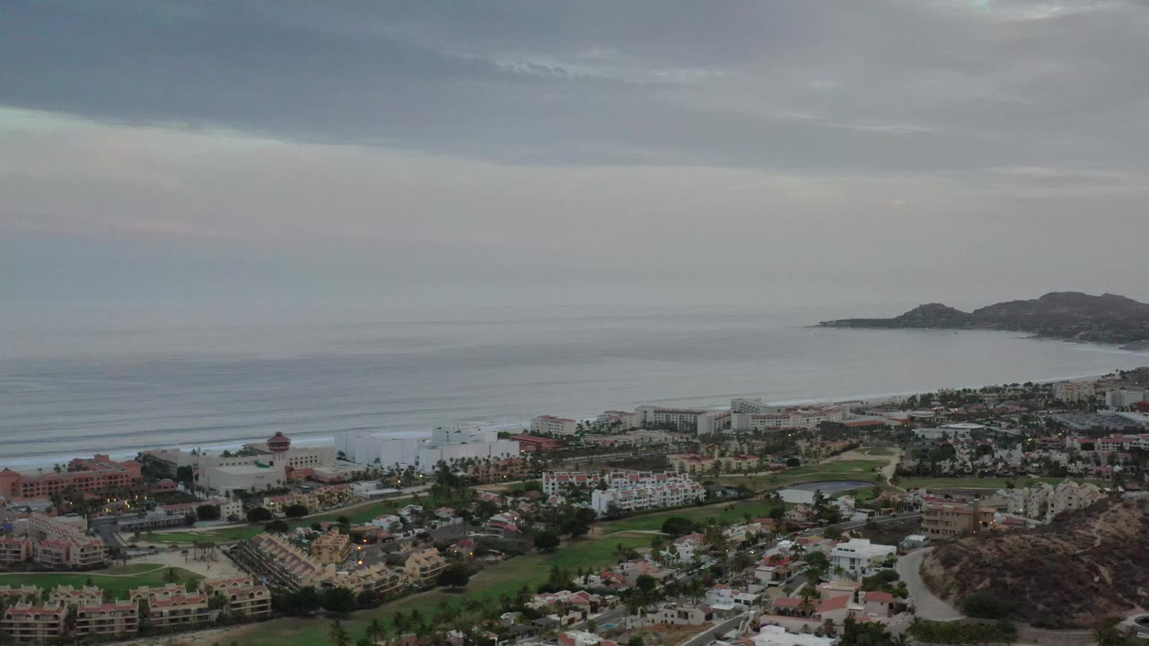 The Los Cabos aerial view holds on dense rooftops, palms, green spaces, shoreline buildings, calm water, and headlands at the right side of the bay, with no visible people.