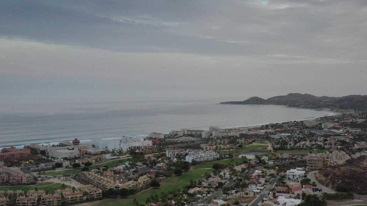 Aerial view of beachfront properties, bay, surf, and hills under gray clouds.