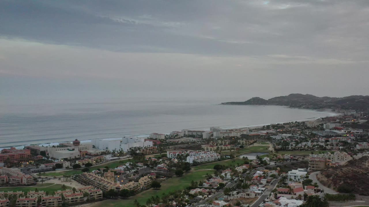High aerial frame of Los Cabos coastline with clustered beachside buildings, a curved bay, low surf, dark hills, and no visible people.