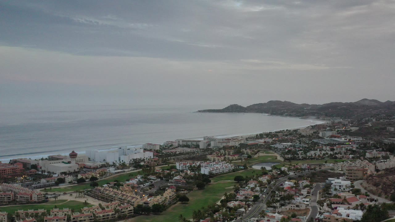 Wide aerial view over Los Cabos, Mexico, showing ocean water, a curving coastline, hillside terrain, roads, homes, green grass areas, and no visible people.