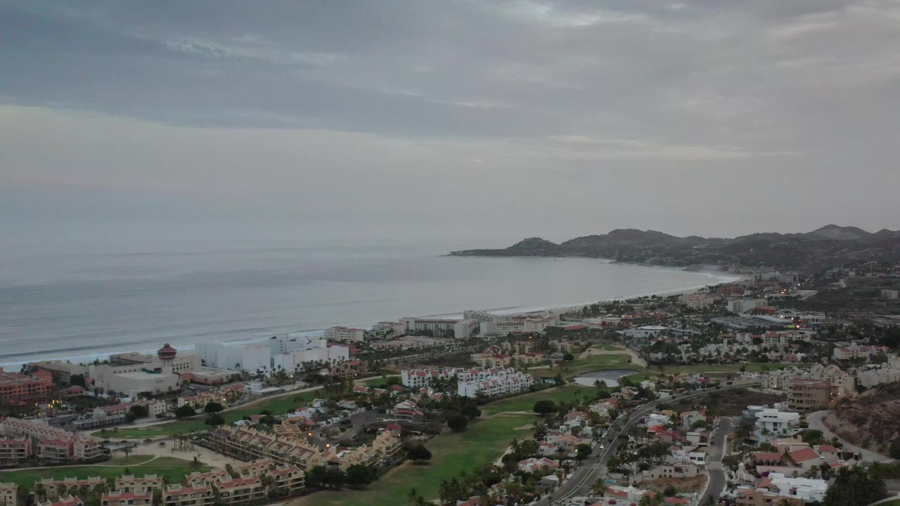 Elevated Los Cabos view with bay water at left, low hills beyond the shoreline, clustered buildings, green fairway-like grass, roads, and no visible people.