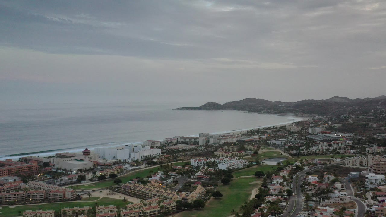 Aerial view of ocean coastline, town roads, buildings, green areas, and hills under a gray sky.