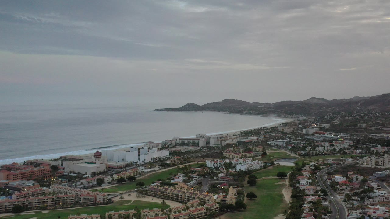 Opening aerial frame of Los Cabos with the ocean on the left, beachfront buildings below, dark hills in the distance, gray cloud cover above, and no visible people.