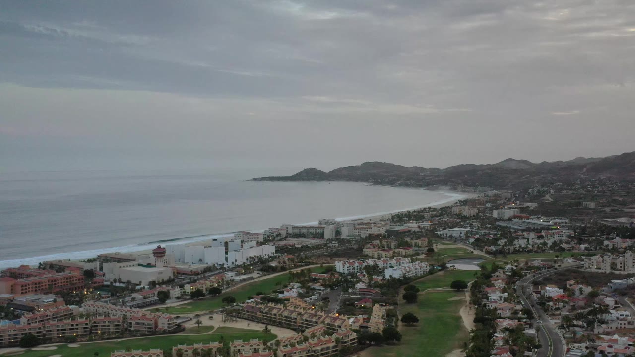 Second aerial frame over Los Cabos showing a broad ocean horizon, white surf along the beach, dense city buildings, green foreground grounds, and no visible people.