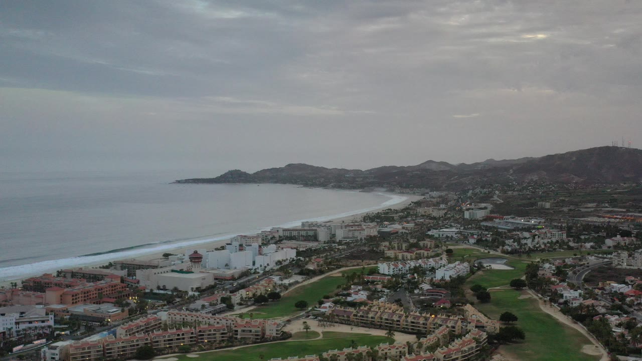 Opening aerial frame over Los Cabos, Mexico, showing ocean and surf at left, developed shoreline, green fairway-like areas, distant mountains, cloudy sky, and no visible people.