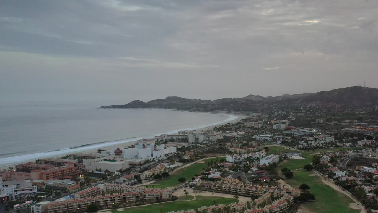 Elevated coastal city view with surf, beach, low mountains, roads, and cloudy sky.