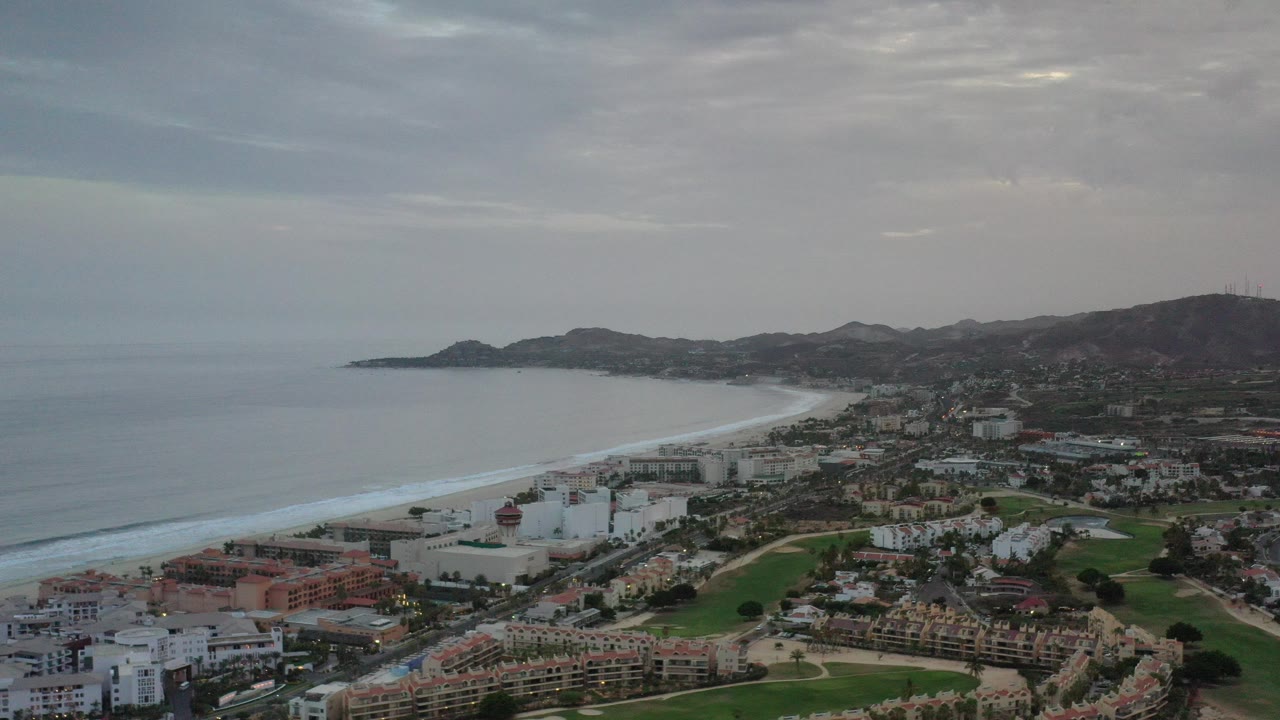 Aerial view of gray ocean, surf, coastal buildings, green fairways, and hills under clouds.